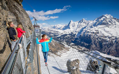 Visitors on a cliffside walkway with snowy Alps view at Schilthorn, Switzerland.