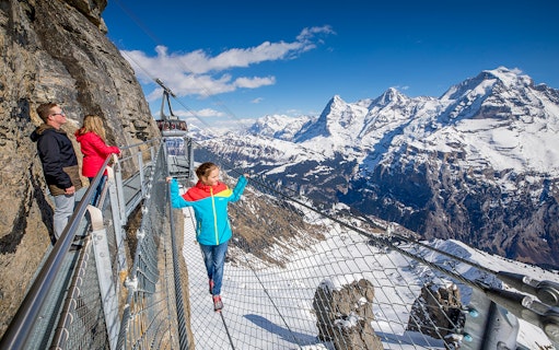 Visitors on a cliffside walkway with snowy Alps view at Schilthorn, Switzerland.