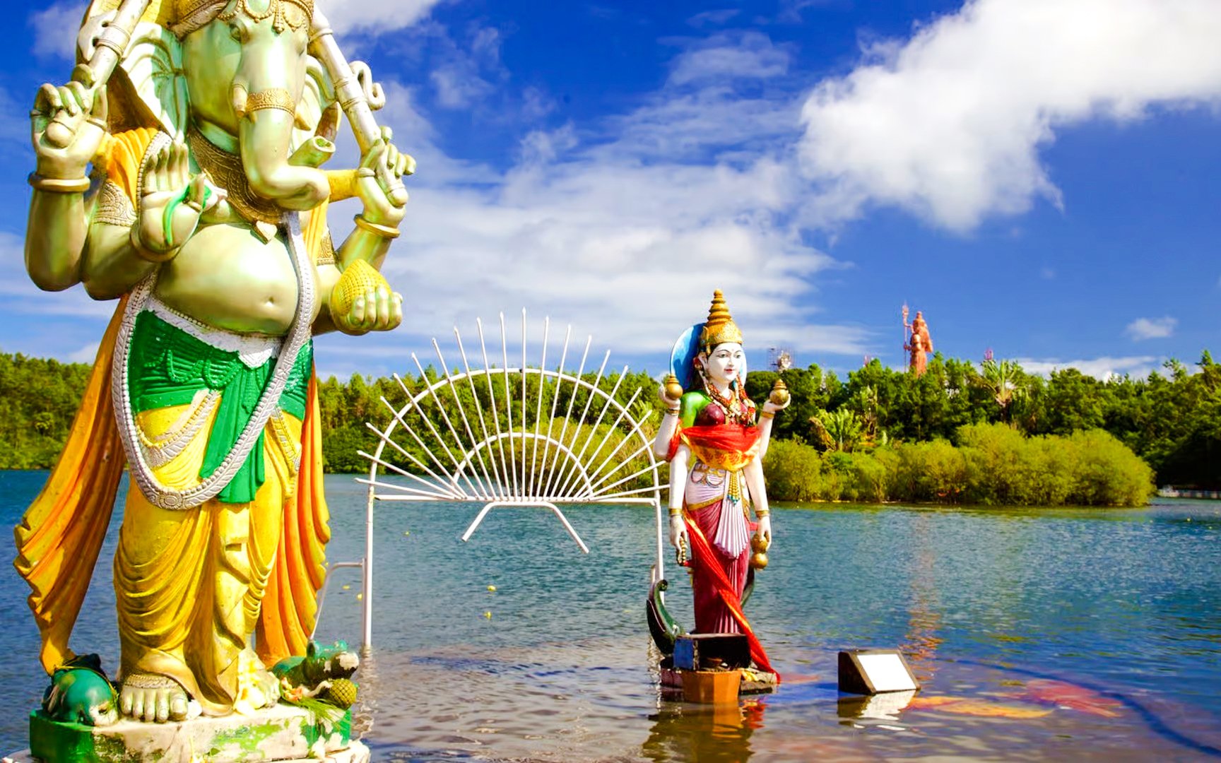Statues of Hindu deities at Ganga Talao, Mauritius, with a lake and lush greenery.