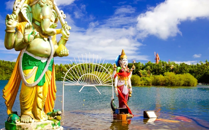 Statues of Hindu deities at Ganga Talao, Mauritius, with a lake and lush greenery.