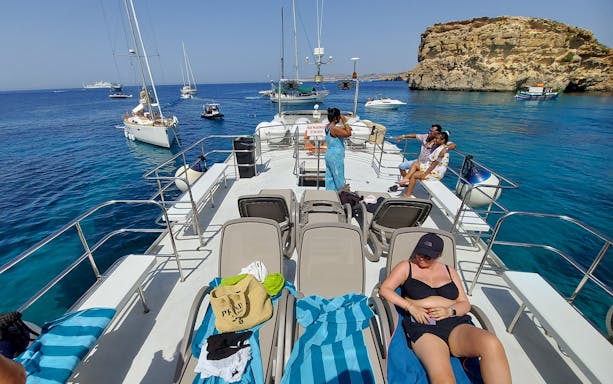 People relaxing on a cruise deck with sun loungers, surrounded by boats and rocky coastline.