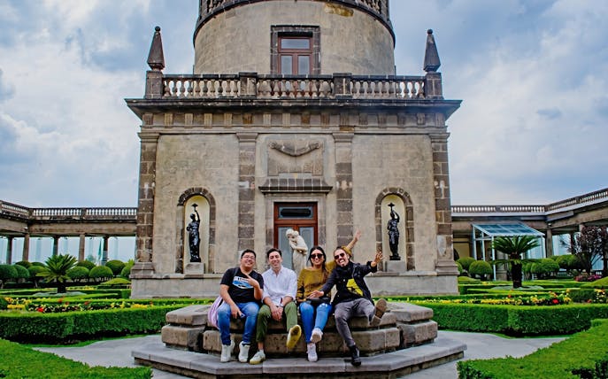 Visitors sitting in front of Chapultepec Castle, Mexico City, with statues and gardens in the background.