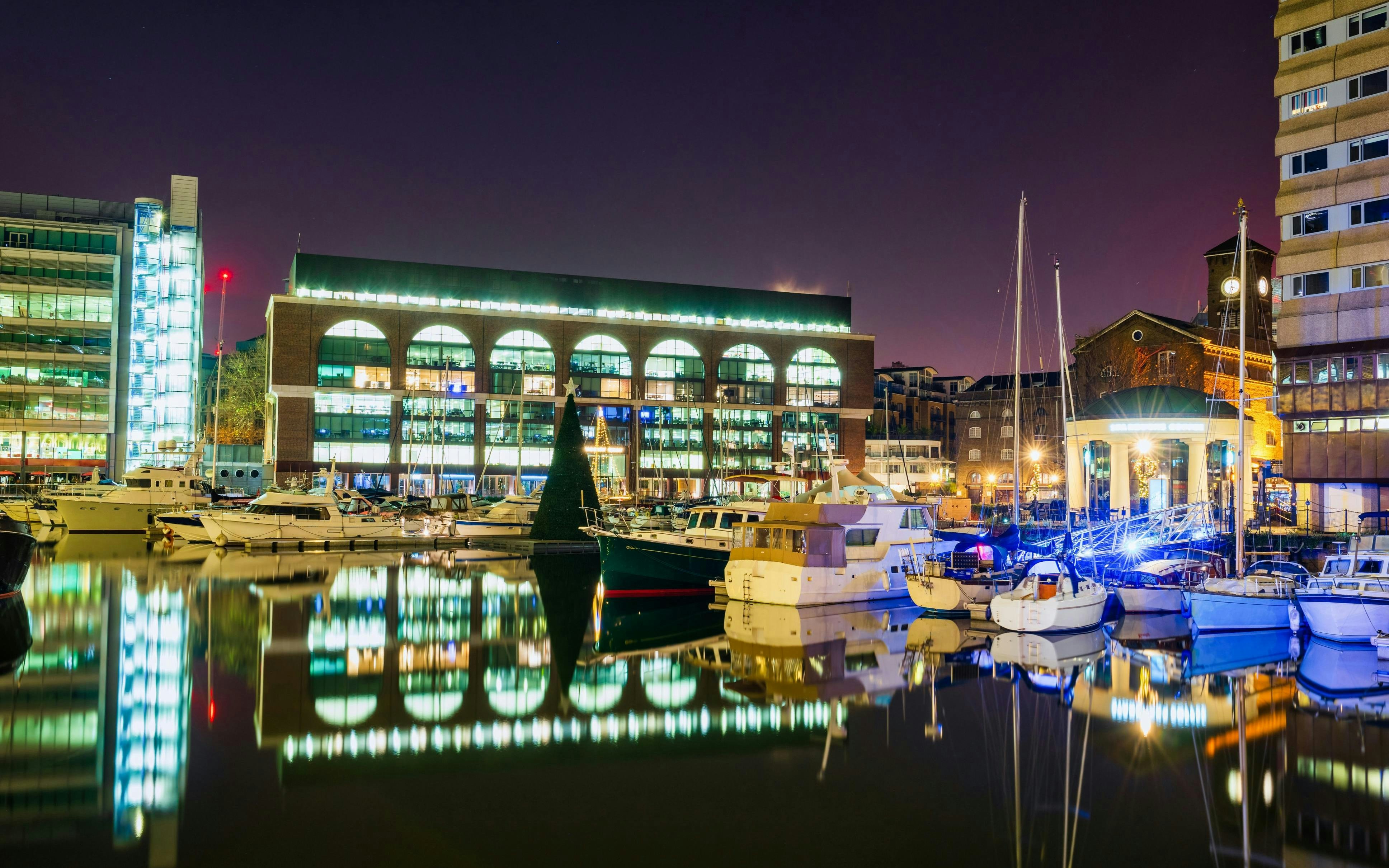 Boats docked at St Katharine Docks in London at night with illuminated buildings.