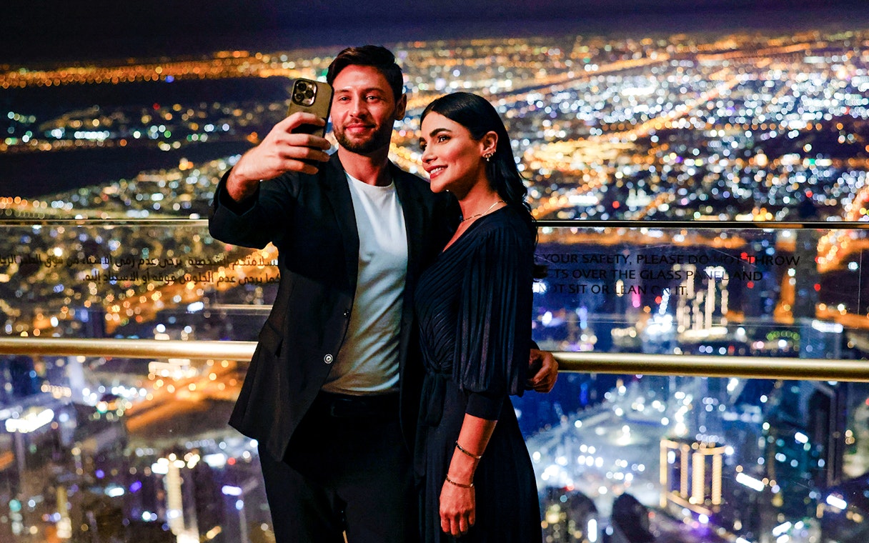 Couple taking selfie at Burj Khalifa Level 152 Lounge with Dubai city lights in background.