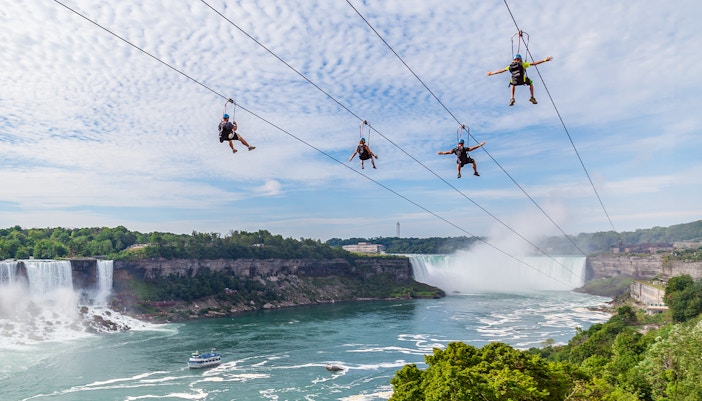 Zip line riders over Niagara Falls with mist and boat below.