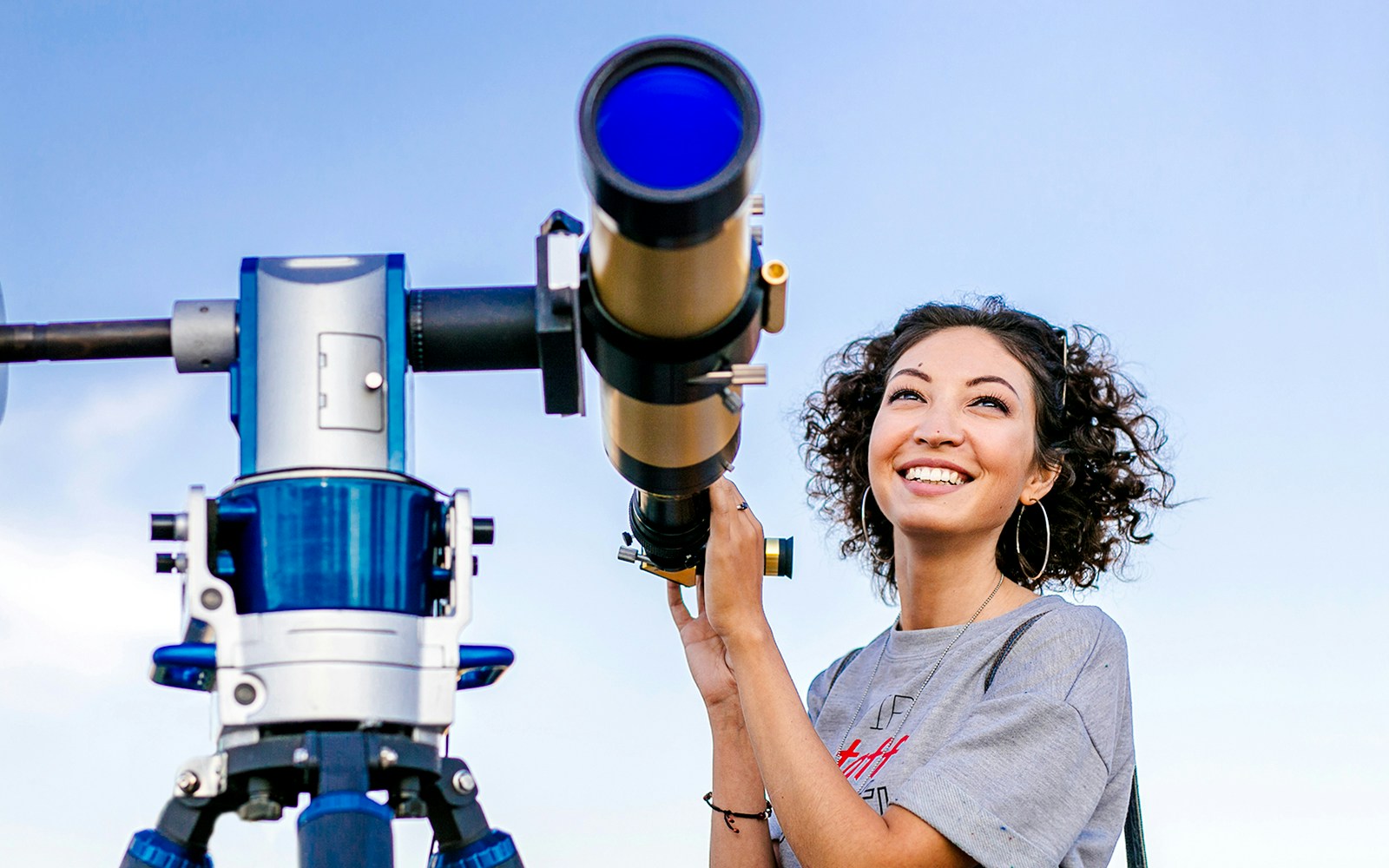 Girl smiling while using a telescope outdoors under a clear blue sky.