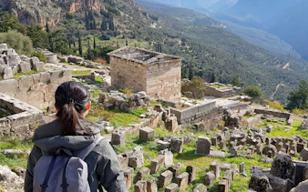 Visitor exploring ancient ruins at Delphi, Greece, with mountainous landscape in the background.