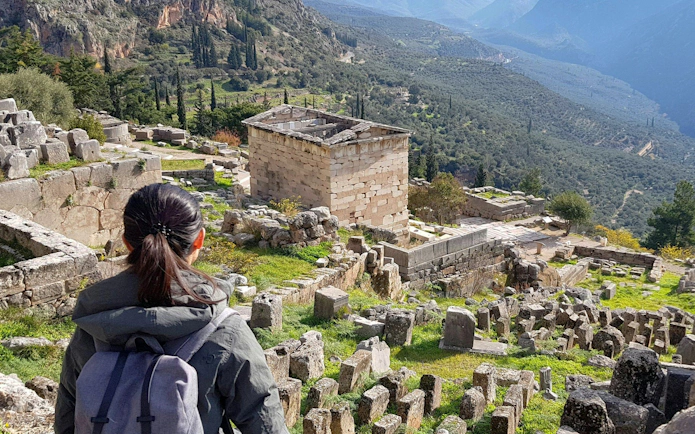 Visitor exploring ancient ruins at Delphi, Greece, with mountainous landscape in the background.