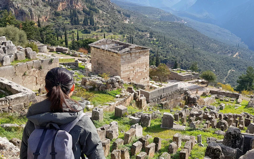 Visitor exploring ancient ruins at Delphi, Greece, with mountainous landscape in the background.
