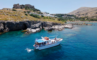 Boat cruising near Lindos cliffs with turquoise sea water.