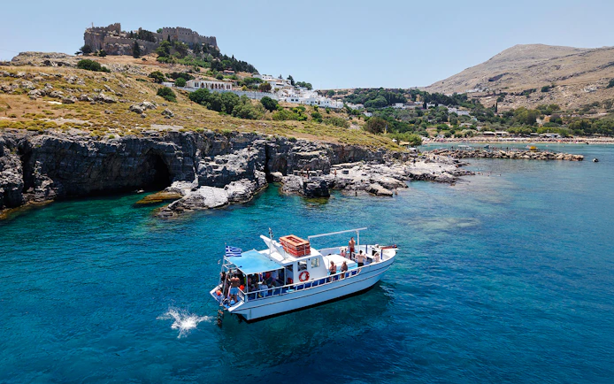 Boat cruising near Lindos cliffs with turquoise sea water.