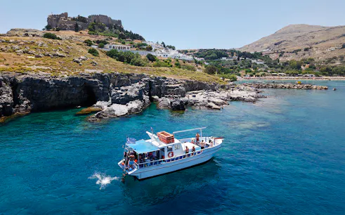 Boat cruising near Lindos cliffs with turquoise sea water.
