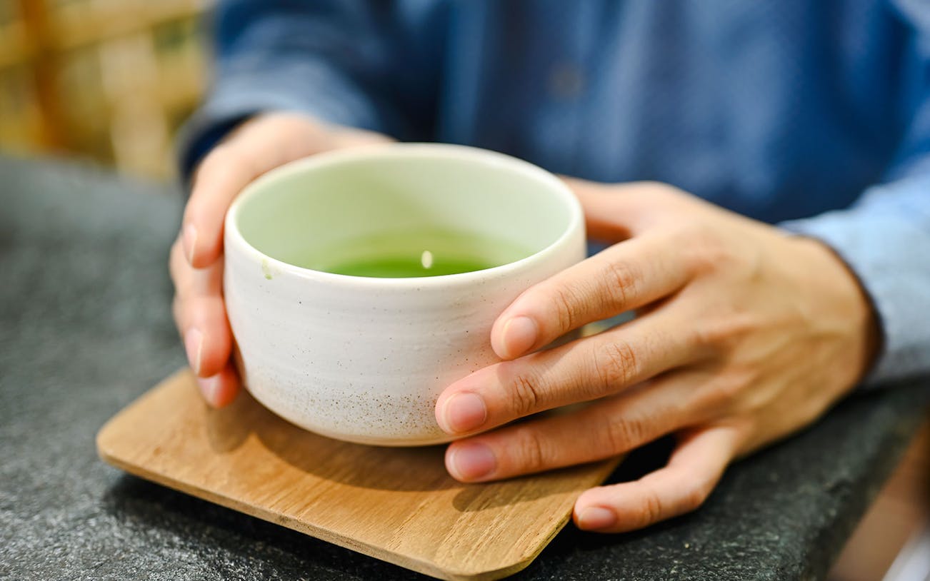 Man holding a bowl of matcha tea on a wooden coaster.
