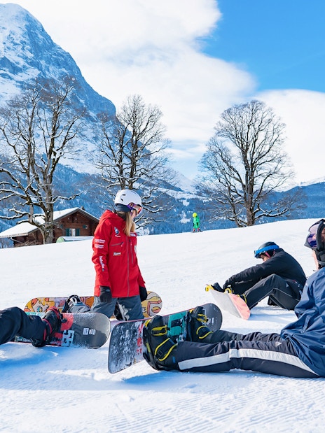 Beginner snowboarders resting on a snowy slope in Grindelwald with scenic mountain views.