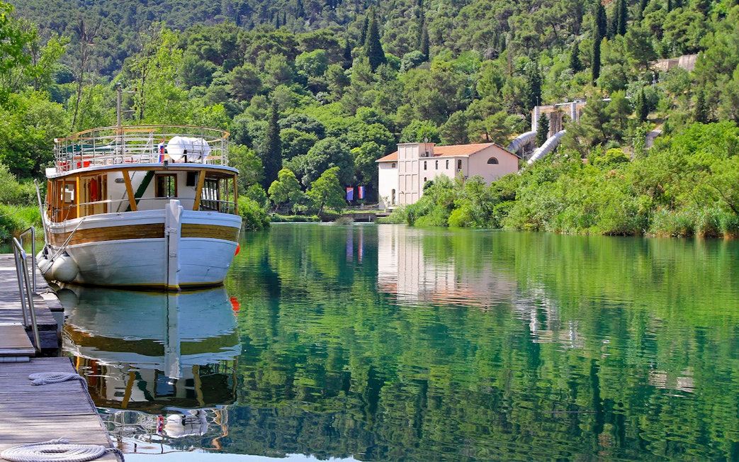 Boat docked on Krka National Park river with lush greenery and historic building in background.