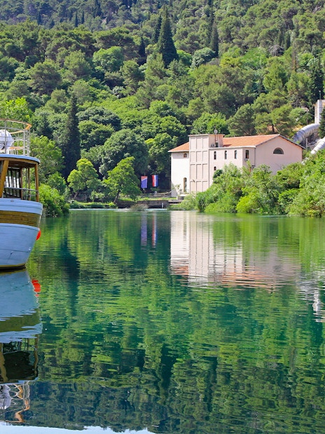 Boat docked on Krka National Park river with lush greenery and historic building in background.