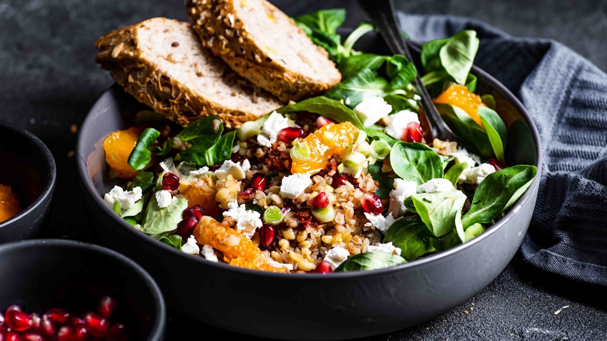 Vegan salad with spinach, pomegranate, orange slices, and bread in a bowl.