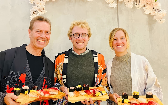 Tourists holding sushi platters, smiling in traditional attire under cherry blossoms.
