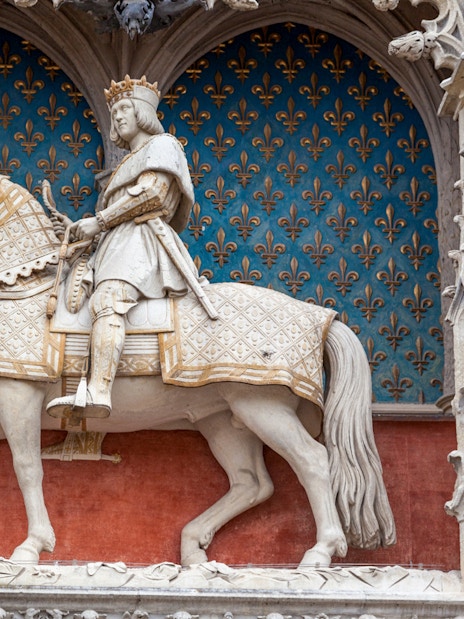 Equestrian statue of a king on a horse with ornate armor, set against a fleur-de-lis backdrop.