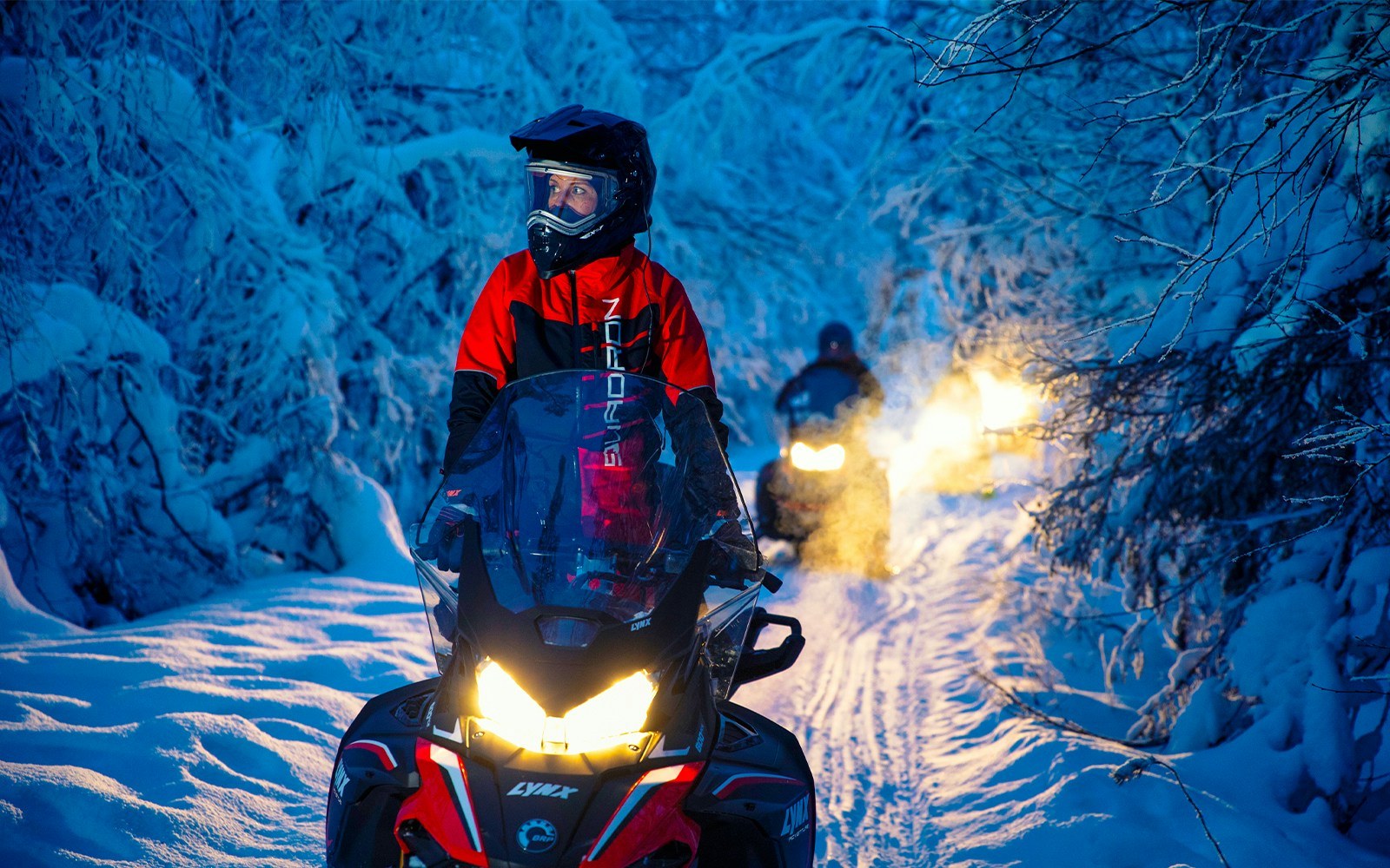 Snowmobiles driving through a snowy forest trail at dusk.