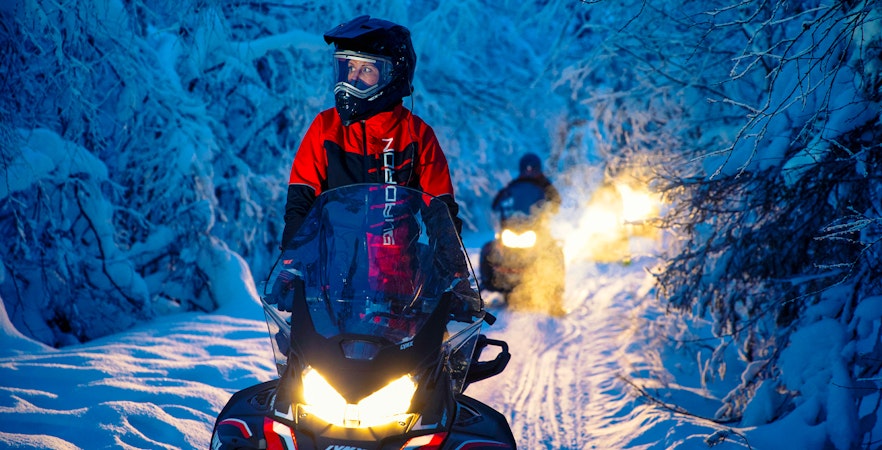 Snowmobiles driving through a snowy forest trail at dusk.