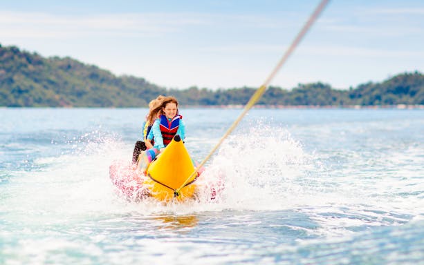 Kids enjoying a banana boat ride on the ocean with distant hills in the background.