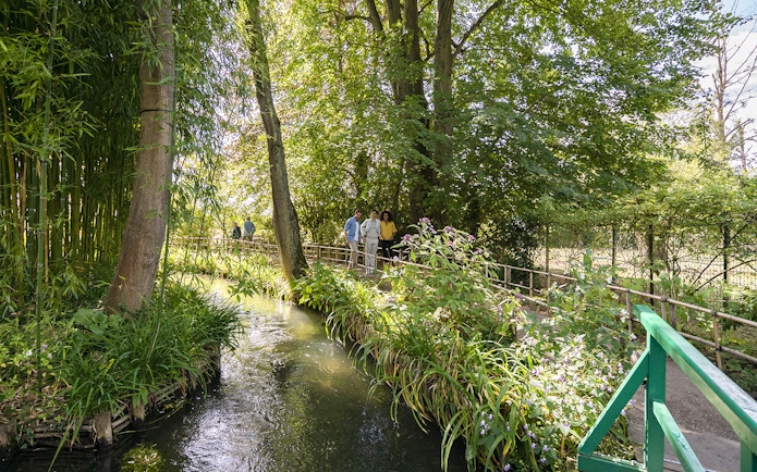 Visitors walking along a path in Giverny Gardens, surrounded by lush greenery and a small stream.