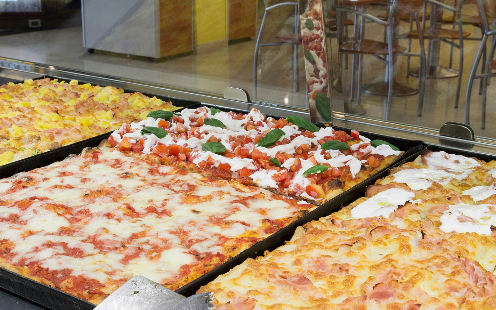 Pizza al taglio varieties with toppings like tomato, basil, and cheese in a display case.