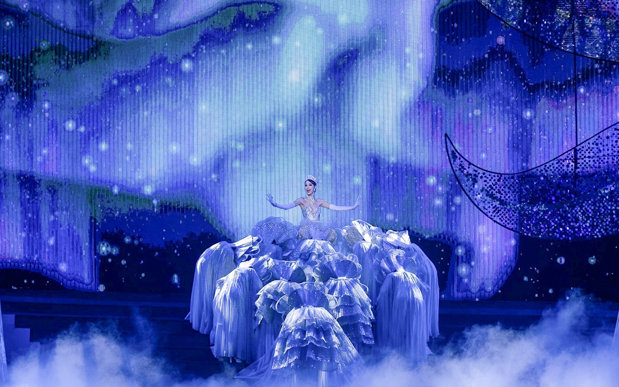 Performer in elaborate costume on stage at Tiffany's Show, surrounded by dancers in blue lighting.