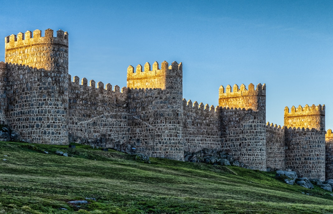 Ancient stone city wall in Ávila, Spain, with sunlit towers and grassy foreground.