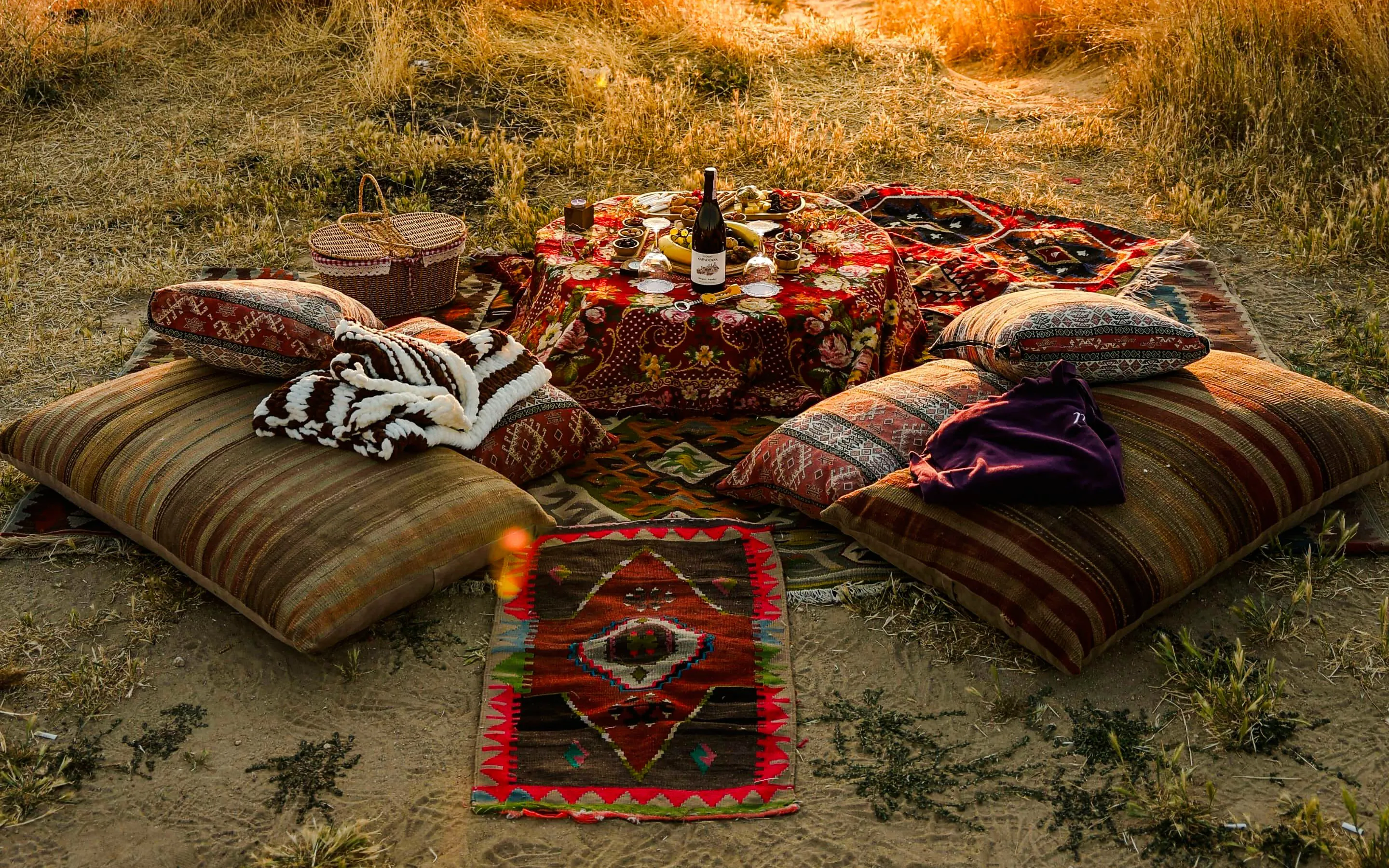 Cushioned picnic setup with wine and snacks in a grassy field, Cappadocia sunset.