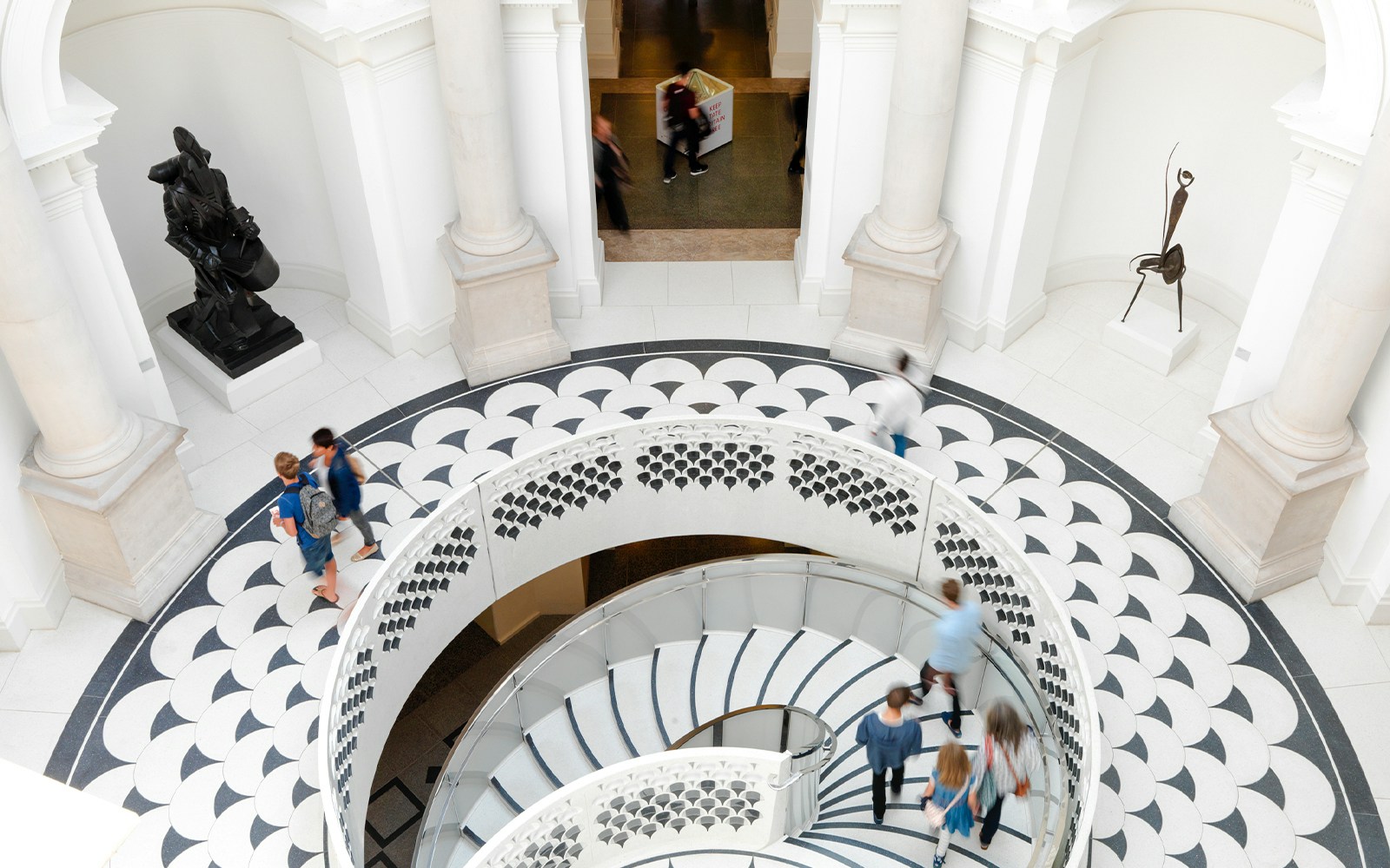 Visitors walking on spiral staircase in Tate Britain interior, London, with sculptures and patterned floor.