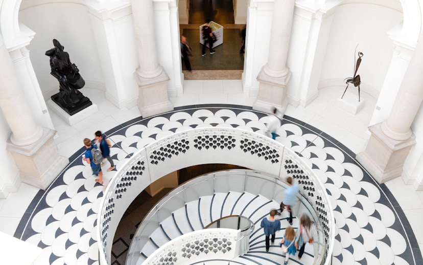 Visitors walking on spiral staircase in Tate Britain interior, London, with sculptures and patterned floor.