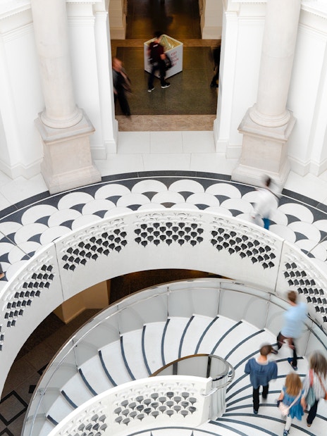 Visitors walking on spiral staircase in Tate Britain interior, London, with sculptures and patterned floor.