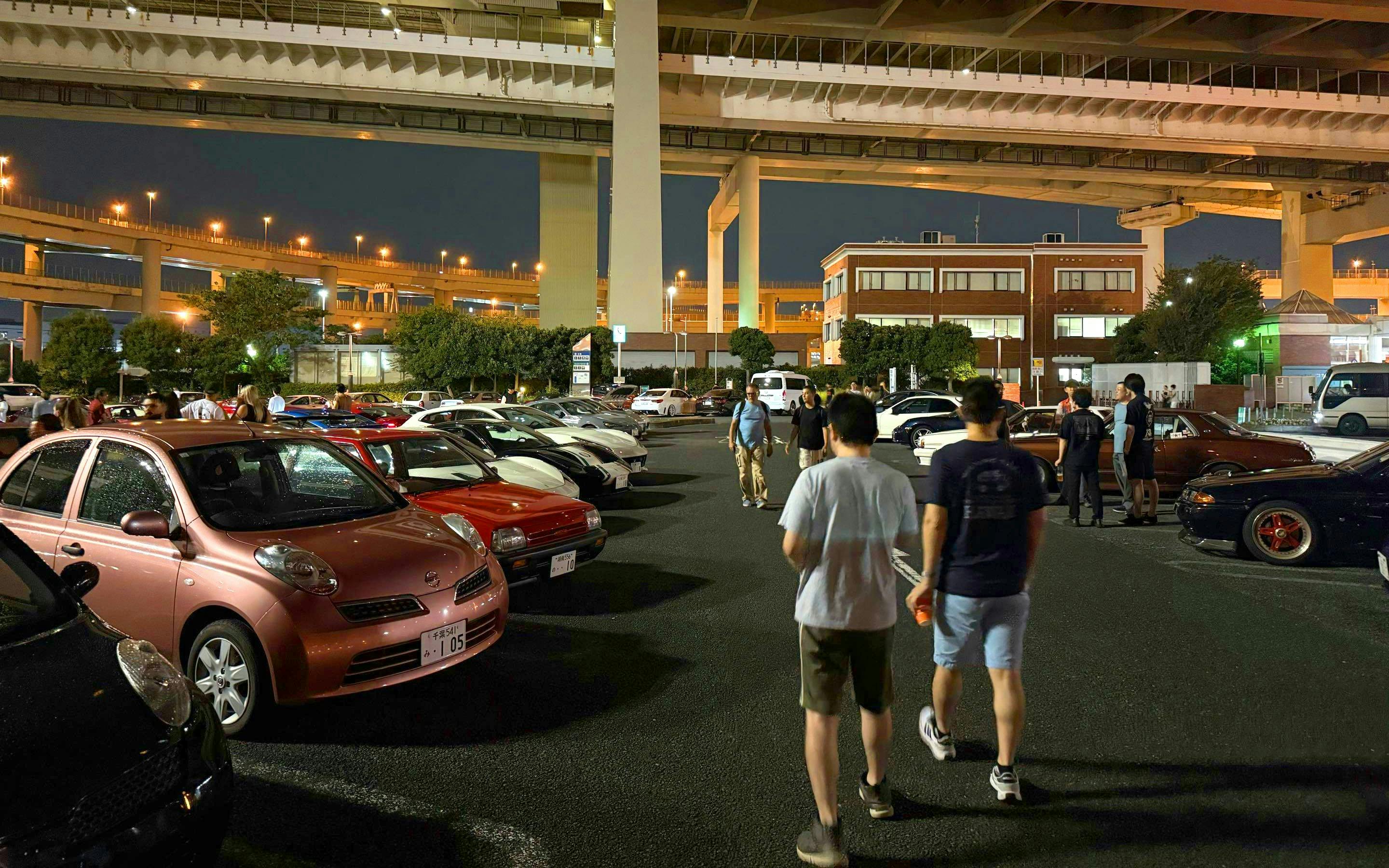 Car enthusiasts gather at Daikoku PA in Tokyo, showcasing diverse vehicles under highway overpasses.