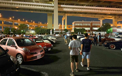 Car enthusiasts gather at Daikoku PA in Tokyo, showcasing diverse vehicles under highway overpasses.
