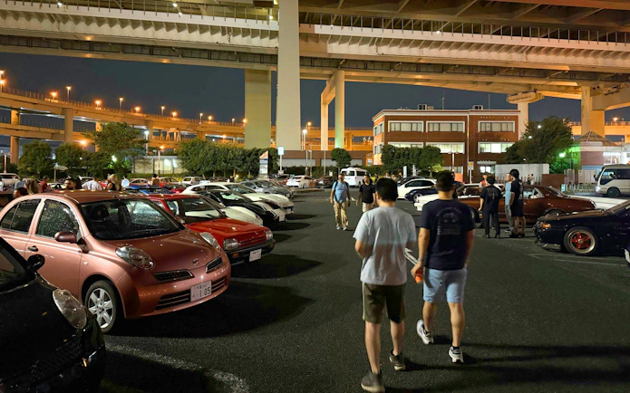 Car enthusiasts gather at Daikoku PA in Tokyo, showcasing diverse vehicles under highway overpasses.