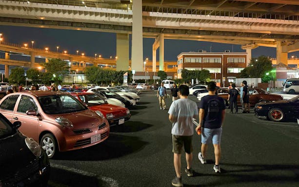 Car enthusiasts gather at Daikoku PA in Tokyo, showcasing diverse vehicles under highway overpasses.