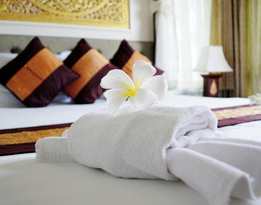 The image shows a neatly made hotel bed with a towel folded into a decorative shape, possibly a swan or similar design, adorned with a white flower.