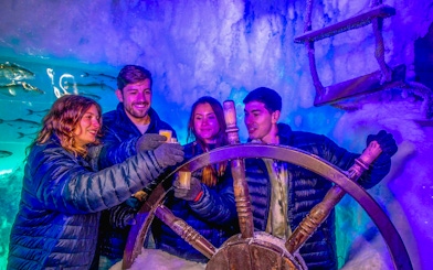 Visitors enjoying drinks at Icebar Amsterdam, holding glasses near an icy ship wheel.