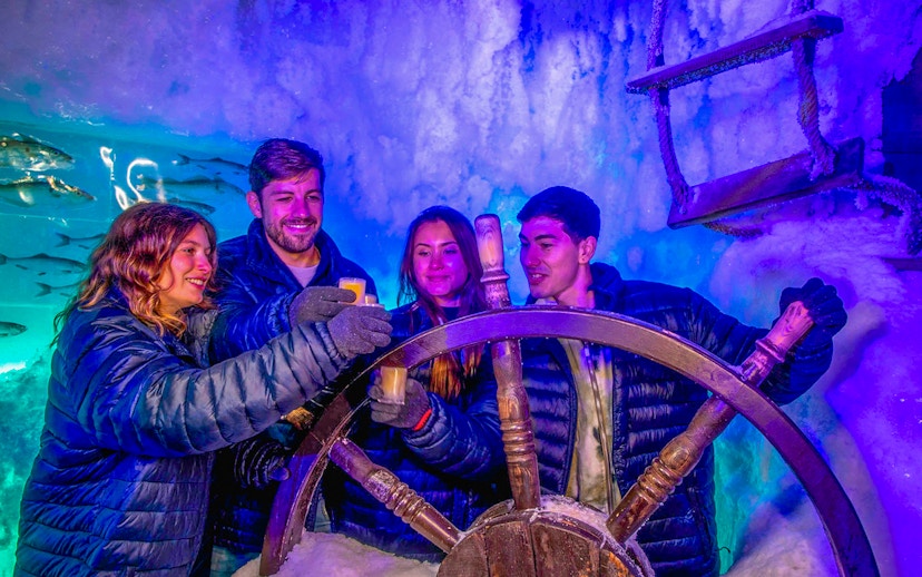 Visitors enjoying drinks at Icebar Amsterdam, holding glasses near an icy ship wheel.