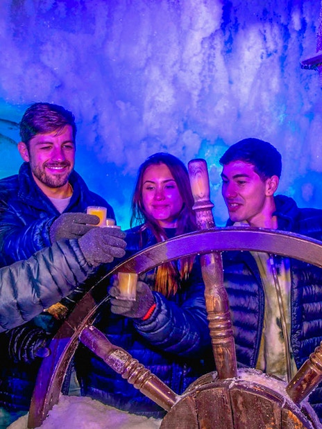 Visitors enjoying drinks at Icebar Amsterdam, holding glasses near an icy ship wheel.