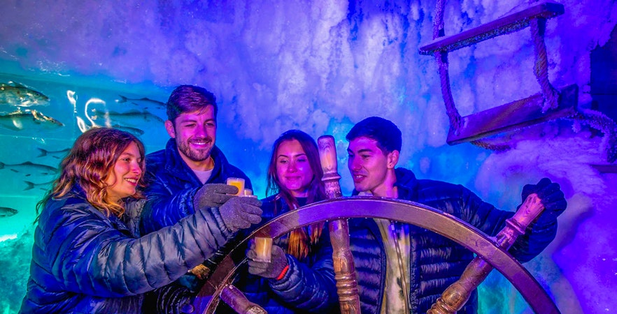 Visitors enjoying drinks at Icebar Amsterdam, holding glasses near an icy ship wheel.