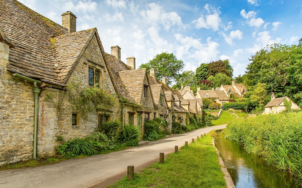 Cotswolds stone cottages along a narrow road with lush greenery, part of a guided day tour from London.