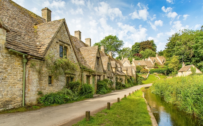 Cotswolds stone cottages along a narrow road with lush greenery, part of a guided day tour from London.