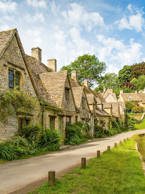 Cotswolds stone cottages along a narrow road with lush greenery, part of a guided day tour from London.