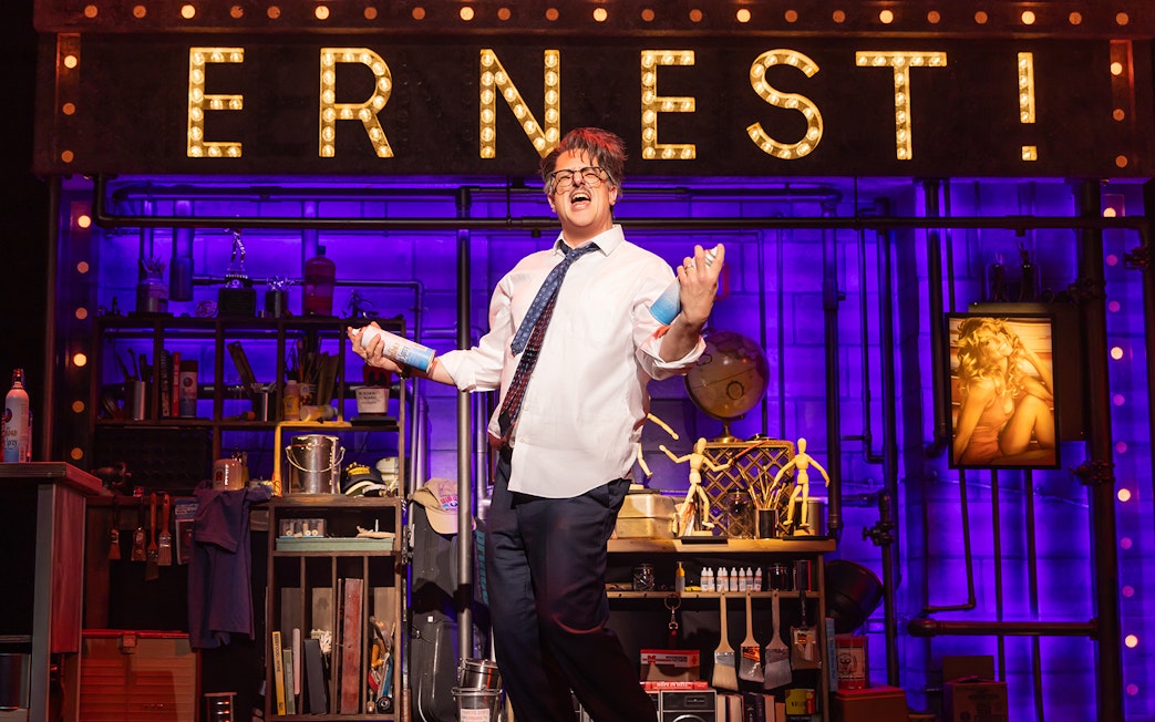 Performer on stage with "ERNEST!" sign, holding spray cans, in a theatrical set for "Death Becomes Her.