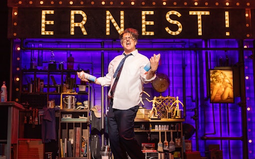 Performer on stage with "ERNEST!" sign, holding spray cans, in a theatrical set for "Death Becomes Her.