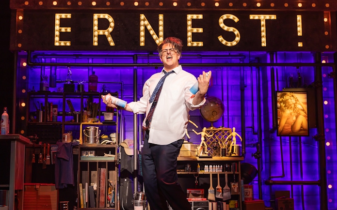 Performer on stage with "ERNEST!" sign, holding spray cans, in a theatrical set for "Death Becomes Her.