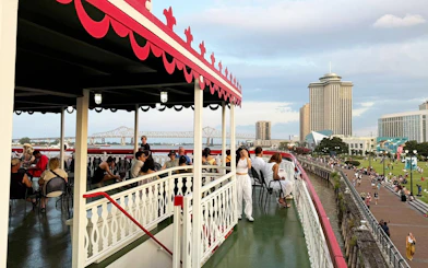People enjoying a jazz cruise on a riverboat in New Orleans with city skyline and bridge in view.